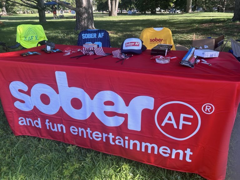 Sober AF Entertainment merchandise table outdoors with branded t-shirts, caps, and flags on a red tablecloth